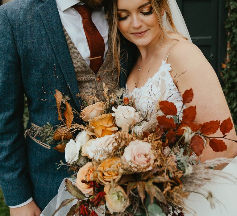 Groom in a blue suit with a brown waistcoat and burnt orange tie with gold tie clip with the bride as she leans against him carrying an autumnal bouquet in sequin Blush by Hayley Paige wedding dress