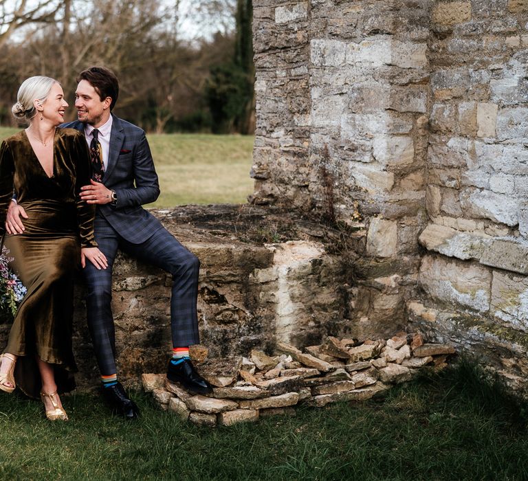 Groom wears colourful socks and sits beside his bride on stone wall outdoors at Bradwell Abbey for relaxed wedding day