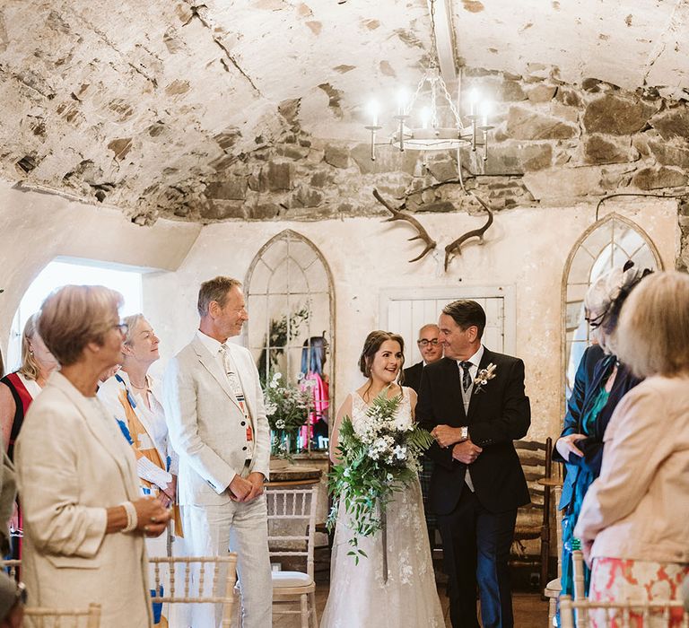 Bride holds oversized bridal bouquet and wears lace wedding dress as she walks down the aisle with her father at Neidpath Castle 