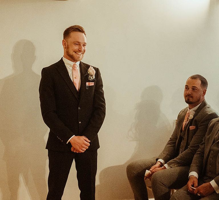 Groom in suit with a oink rose buttonhole and pink tie waits at the altar for the bride 