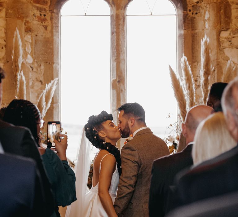 Bride & groom kiss in front of large window at Pentney Abbey surrounded by pampas grass arrangements with Autumnal colour scheme 