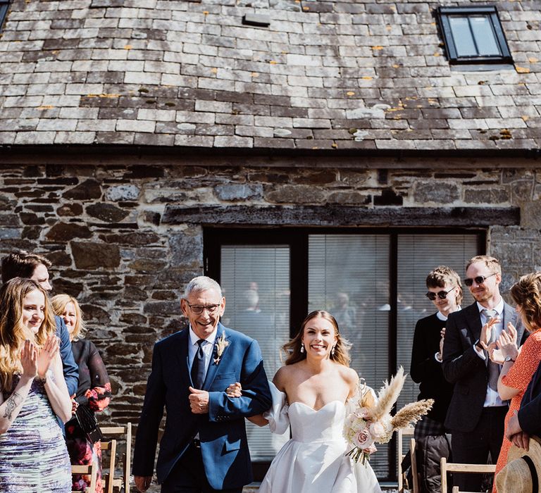 Bride in strapless wedding dress with puff sleeves is walked down the aisle by her father for the outdoor ceremony 