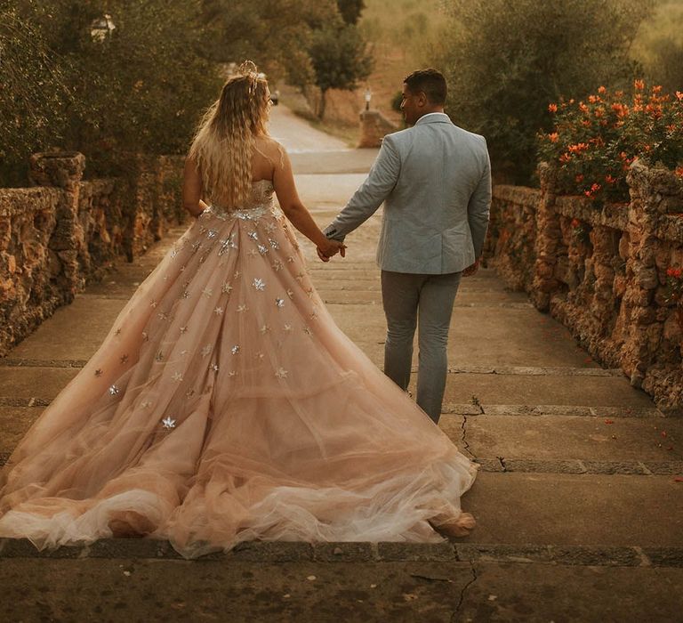 Bride wears celestial wedding for Mallorca wedding whilst walking outdoors with her groom in dusty blue suit 