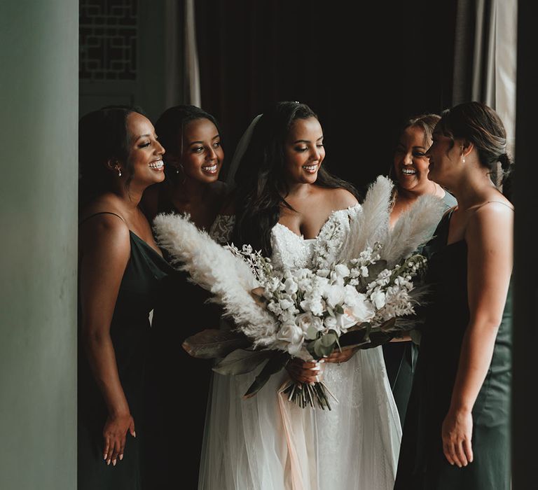 Bride holds oversized pampas grass bouquet and stands with her bridesmaids who wear green cowl neck midi dresses 