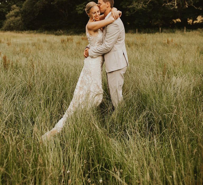 Groom in a light wedding suit hugs the bride as he kisses her on the forehead for their couple portraits 