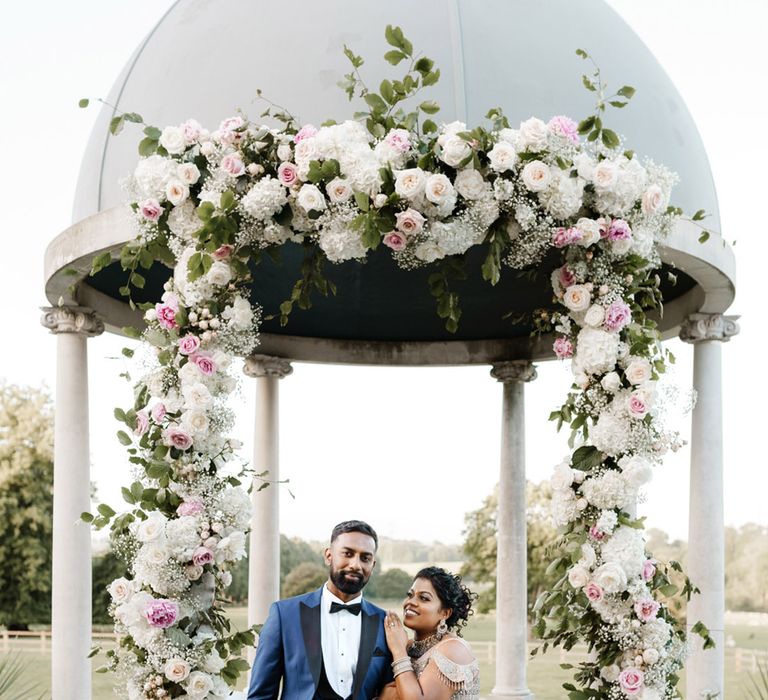 Bride wears gold embellished traditional Sri Lankan wear complete with statement jewellery as she stands beside her groom in black tie and blue suit