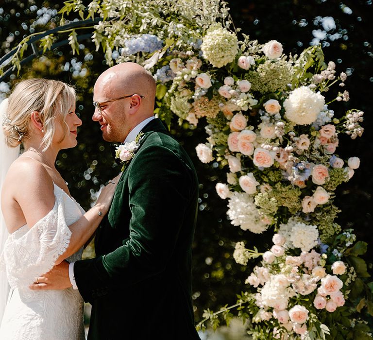 Bride wears lace off the shoulder wedding dress whilst stood with her groom in green velvet black tie 