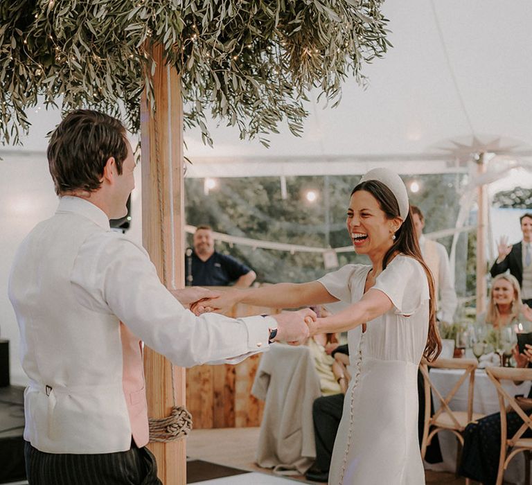 Bride in satin wedding dress and chunky pearl headband with the groom in a pink waistcoat share their first dance together 