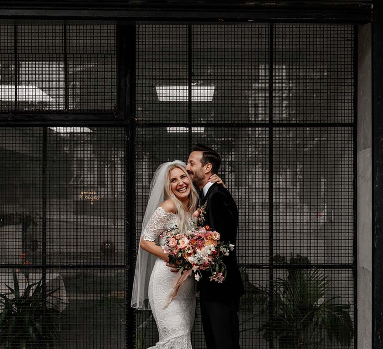 Bride in boho two piece lace bridal outfit with large colourful mixed bouquet and groom in black tux with velvet look bow tie embracing at Hackney Studios