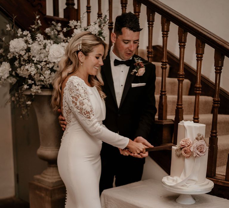 The bride and groom cut their white iced two tier wedding cake with pink flower decorations 