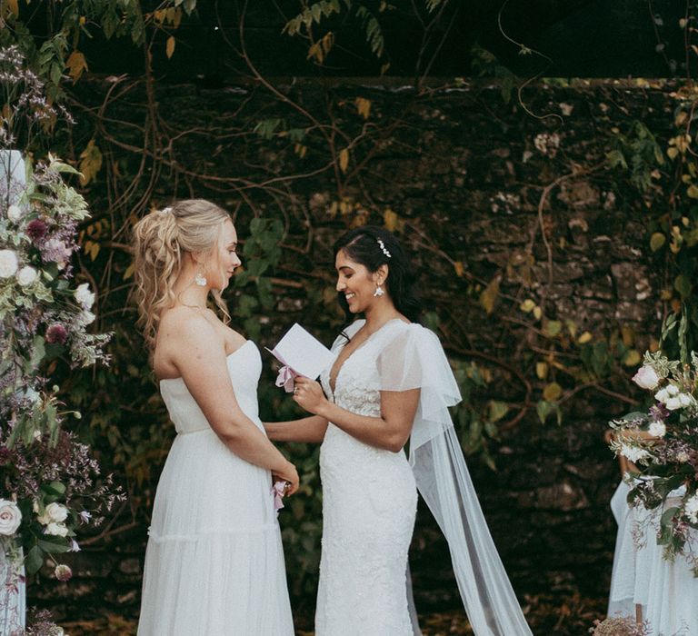 Brides in sleeveless ruched wedding dress and short sleeve v neck wedding dress with puff lace shoulders, reading vows to each other and matching earrings