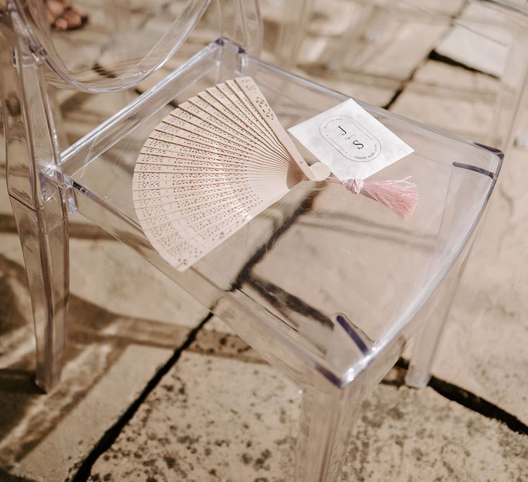 Clear acrylic chairs with biodegradable confetti and wooden fans for guests during the summer weather 