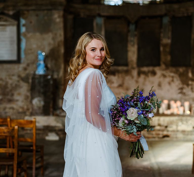 Bride wears her blonde hair in curls and pearl embellished wedding cape