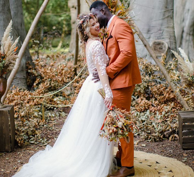 Bride holding dried autumnal wedding bouquet wearing long sleeved v neck soft ivory dress with lace floral detailing on the bodice and sheer sleeves embracing groom in burnt orange rust coloured suit with brown tie, pocket square, brown accessories and dried flower, rose and pampas grass boutonniere  