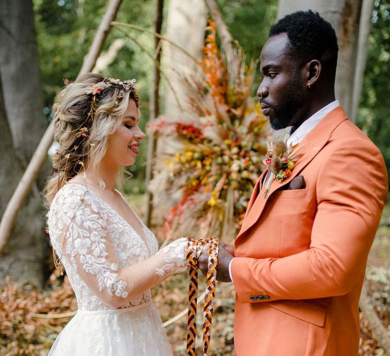 Bride in long sleeved v neck soft ivory dress with botanical embroidered details doing handfesting ceremony with groom in burnt orange rust coloured suit with brown tie, pocket square and brown accessories at a harvest wedding