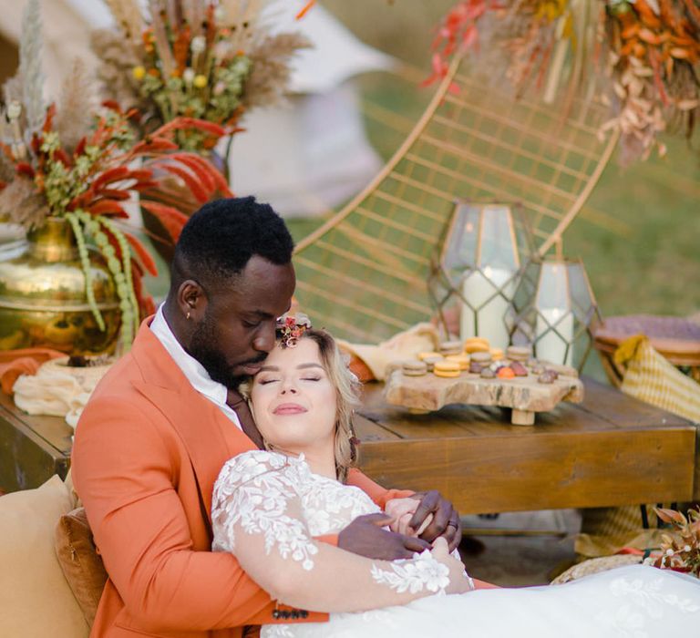 Groom in burnt orange rust coloured suit with brown tie, pocket square, brown accessories and dried flower, rose and pampas grass boutonniere and bride in long sleeved v neck soft ivory dress with lace floral detailing on the bodice and sheer sleeves hugging and relaxing at a harvest wedding with autumnal colours behind them