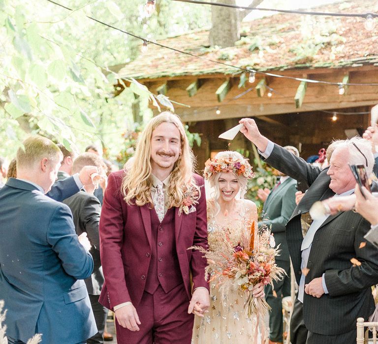 Groom in maroon suit with floral tie and matching pocket square walks alongside his bride in embellished wedding dress with sequin stars