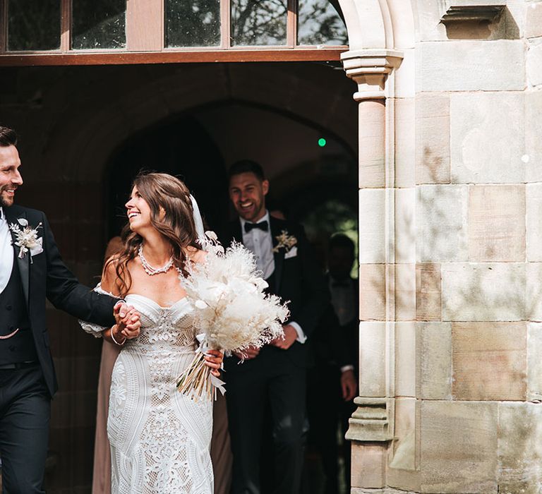 Groom in black tuxedo with gold chain with bride in fitted lace wedding dress and detachable sleeves walking out of the ceremony carrying a dried flower bouquet