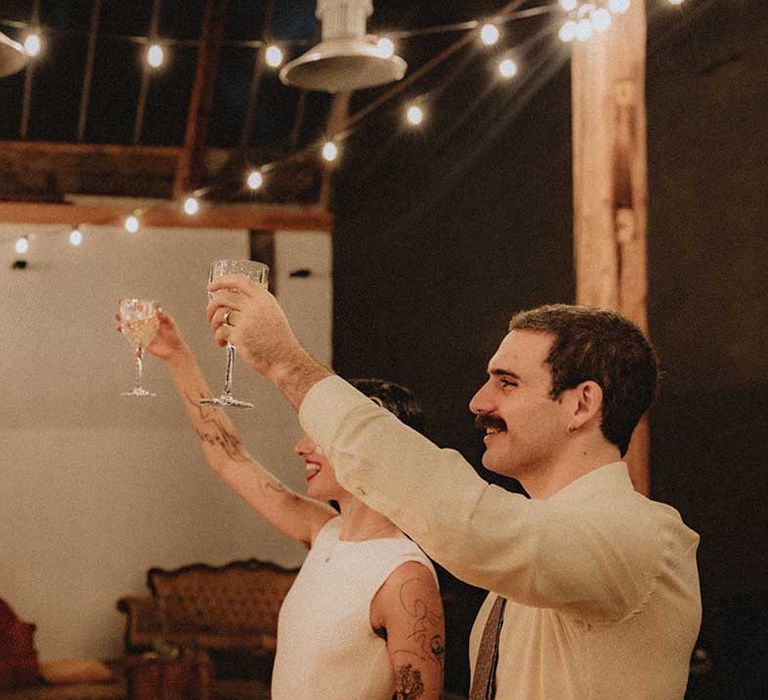 Lisa & Gabriel lift their glasses in cheers during industrial styled reception with festoon lighting 