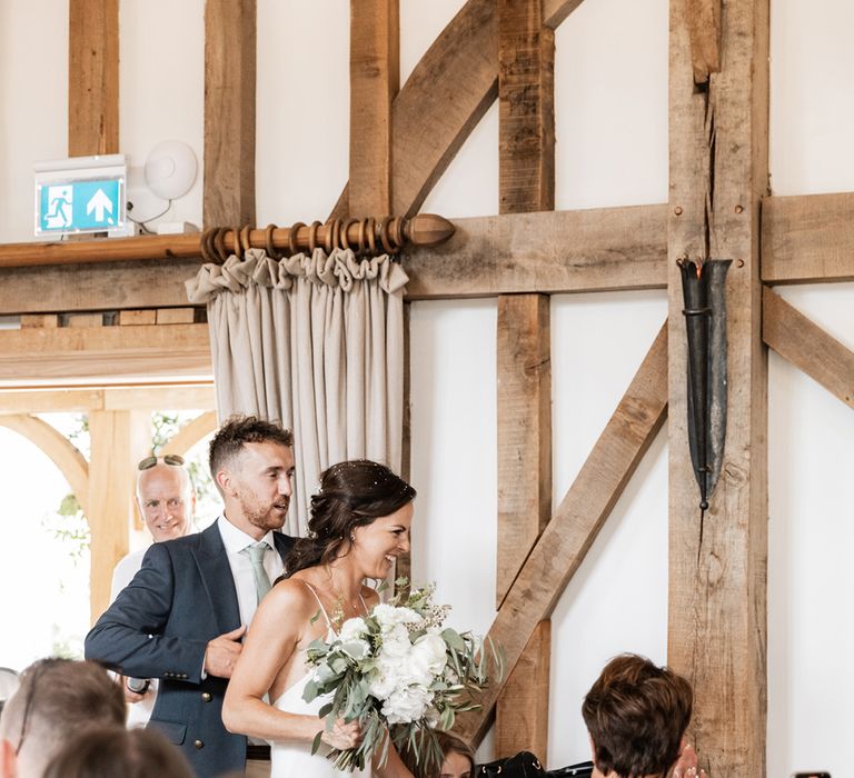 Bride carries white floral bouquet and walks into rustic barn wedding venue for reception 