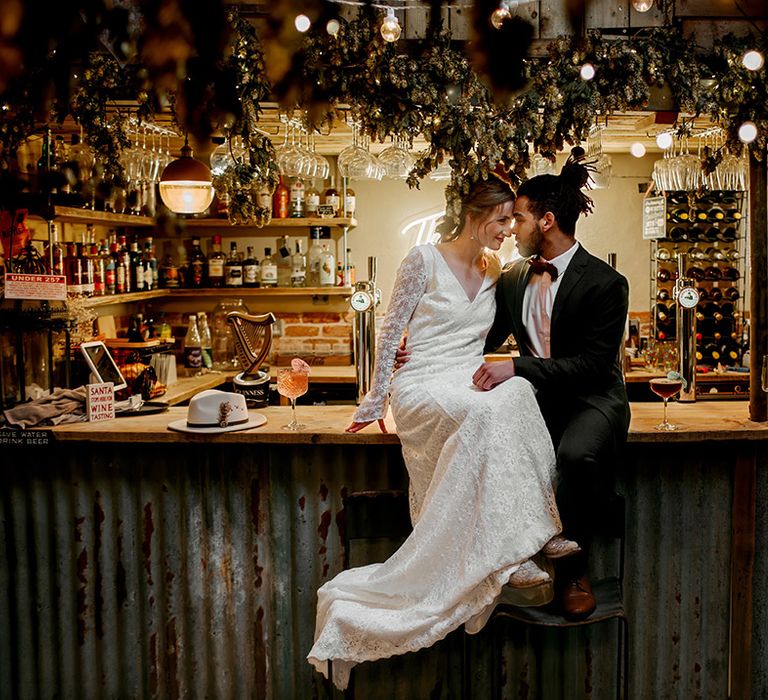 Groom in classic black suit with burgundy bowtie embracing bride in long sleeve lace wedding dress sitting on the bar at barn wedding venue 