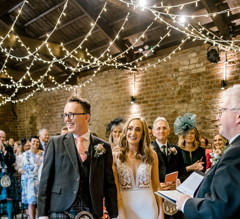 Groom in kilt stands beside his bride in appliqué styled wedding dress with fitted skirt 