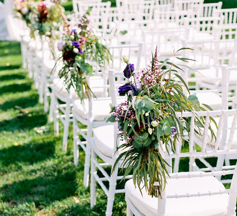 Purple and pink wedding flowers with greenery decorating the end chairs next to the aisle for an outdoor wedding ceremony 