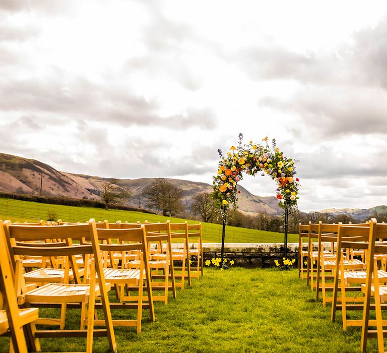 Colourful floral archway to the front of outdoor aisle lined by wooden chairs 