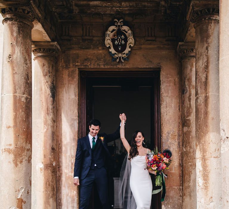 Bride in white satin cowl neck Kyha wedding dress exiting Elmore Court with the groom in a three piece navy suit and green tie 