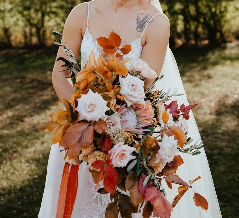 Bride holds burnt orange bridal bouquet with pale pastel roses and Autumnal leaves tied with raw edge orange ribbon 