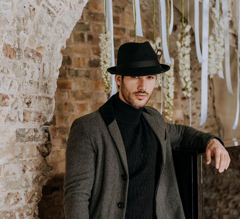 Groom holding bouquet with white roses, white peonies and white chrysanthemums tied with red ribbon wearing dark grey jacket, black turtle neck, black trousers and a black fedora hat 