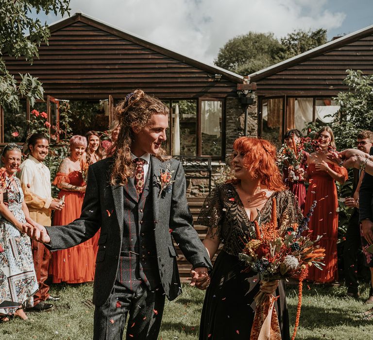 Bride in embellished black wedding dress holding large orange dried flower and eucalyptus bouquet with orange ribbon holding hands with groom in dark grey three piece suit and burgundy flower patterned tie doing orange confetti exit 