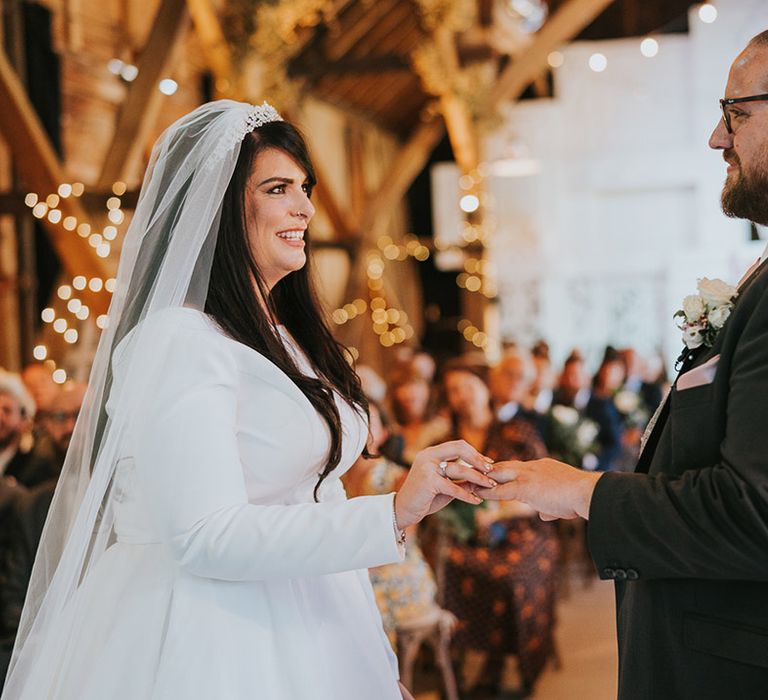 Bride in high neck long sleeve wedding dress with tiara and veil puts the ring on the groom's finger for their wedding ceremony at Preston Court