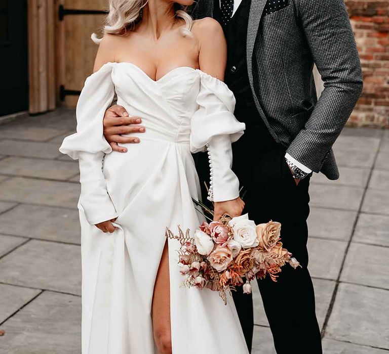 Groom in grey blazer, black waistcoat and navy polkadot tie embracing bride in Nori Ivy off the shoulder long sleeve wedding dress with slight cowl neck and front leg slit 