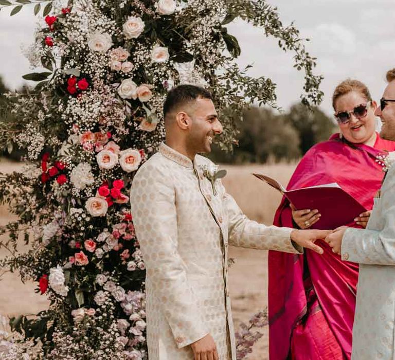 Groom in cream and gold Indian sherwani holding hand of groom in mint green Indian sherwani with wedding celebrant in magenta lehenga standing in front of large garden rose, baby's-breath and foliage floral columns