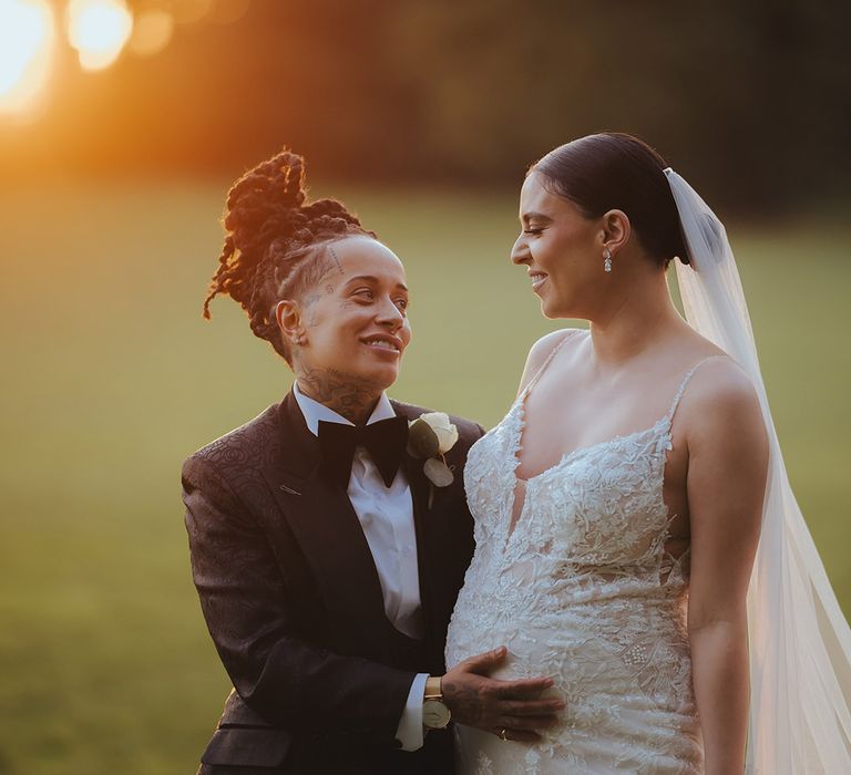 Bride in black snakeskin print tuxedo with black bowtie and white rose boutonniere and golden watch holding bride's baby bump, wearing lace sleeveless v neck Enzoani wedding dress with tulle finish and church length veil at golden hour couples photoshoot 