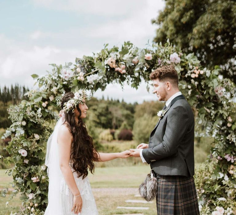 Bride in celestial 3d appliqué sheer overlay wedding dress and bridal flower crown holding hands with groom in dark grey blazer and waistcoat, dark tartan tie with white flower boutonniere, dark grey and blue tartan kilt and wedding sporran at The Barn at Harburn