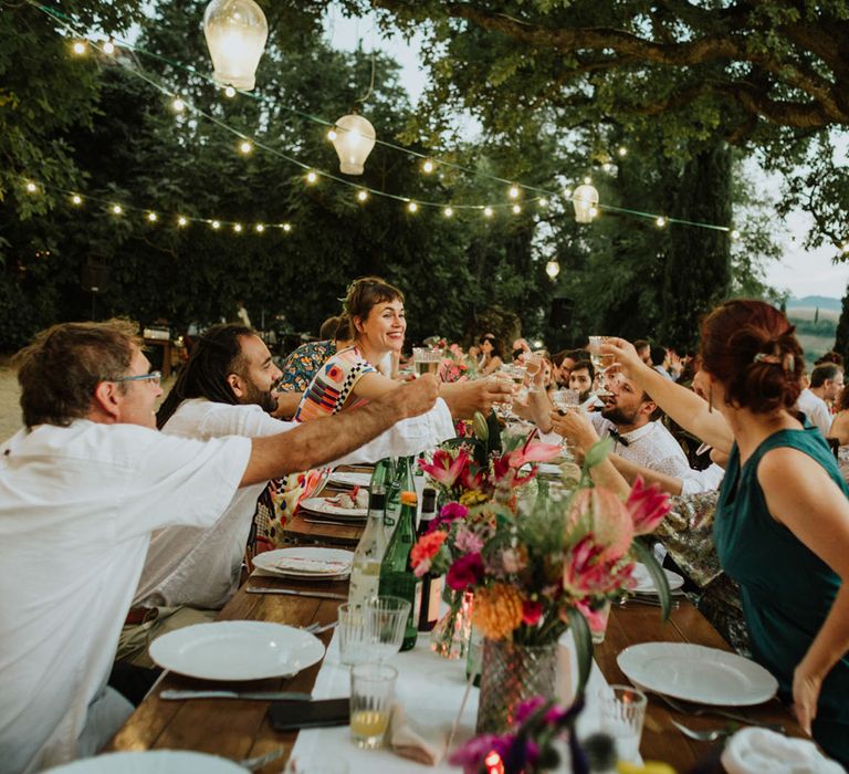 Guests at outdoor wedding in Italy cheers with champagne glasses surrounded by fairylights and pink flower decor