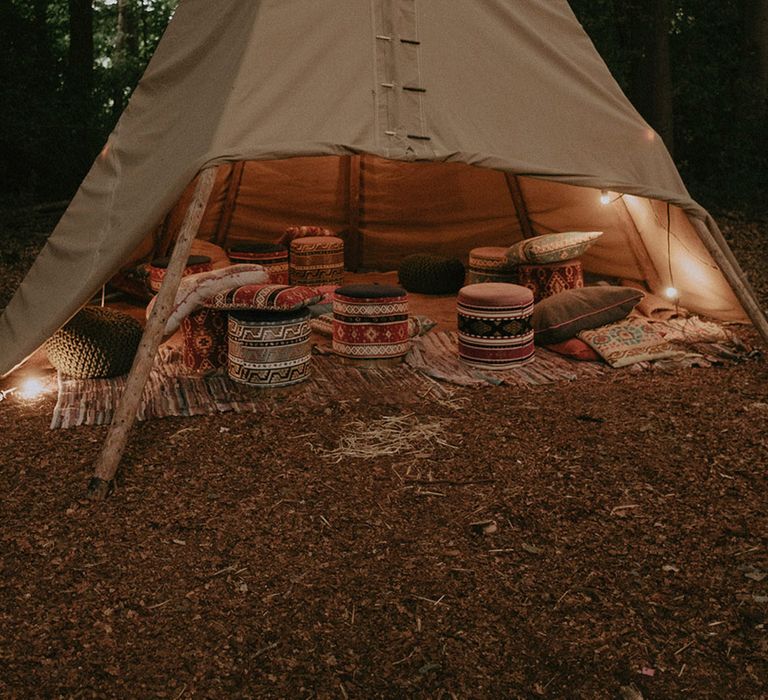 Cosy outdoor tipi area with stools, cushions, and throw blankets at boho wedding 