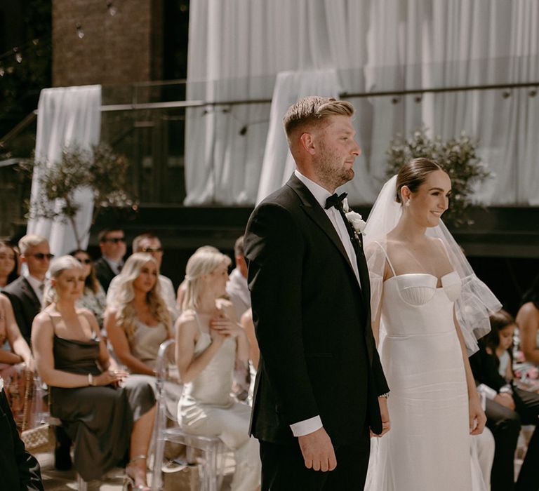 Groom in black tuxedo with white buttonhole stands with the bride for their civil wedding ceremony in London with white flowers and drapery