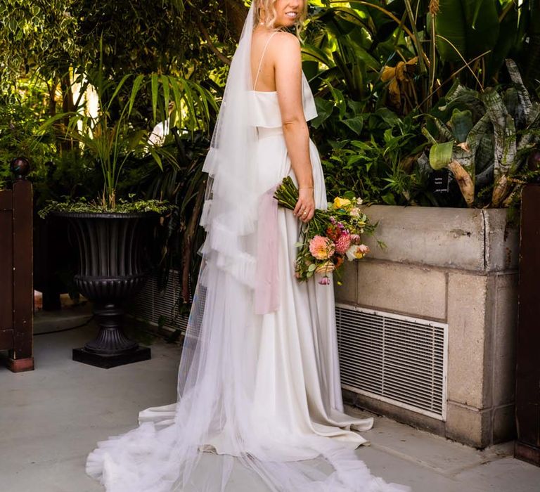 Bride in satin Catherine Deane bridal separates with ruffle wedding veil holding pink and orange bouquet with pink ribbon 