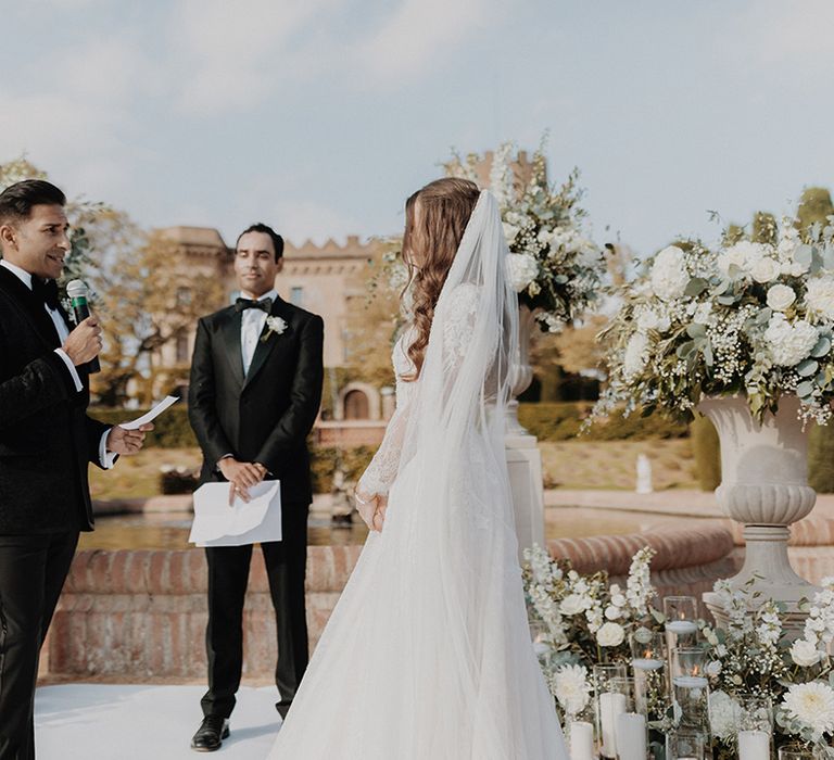 Groom in black tuxedo with black bow tie and white carnation and dried flower boutonniere reading vows to bride wearing lace long sleeve Pronovias wedding dress and church-length veil 