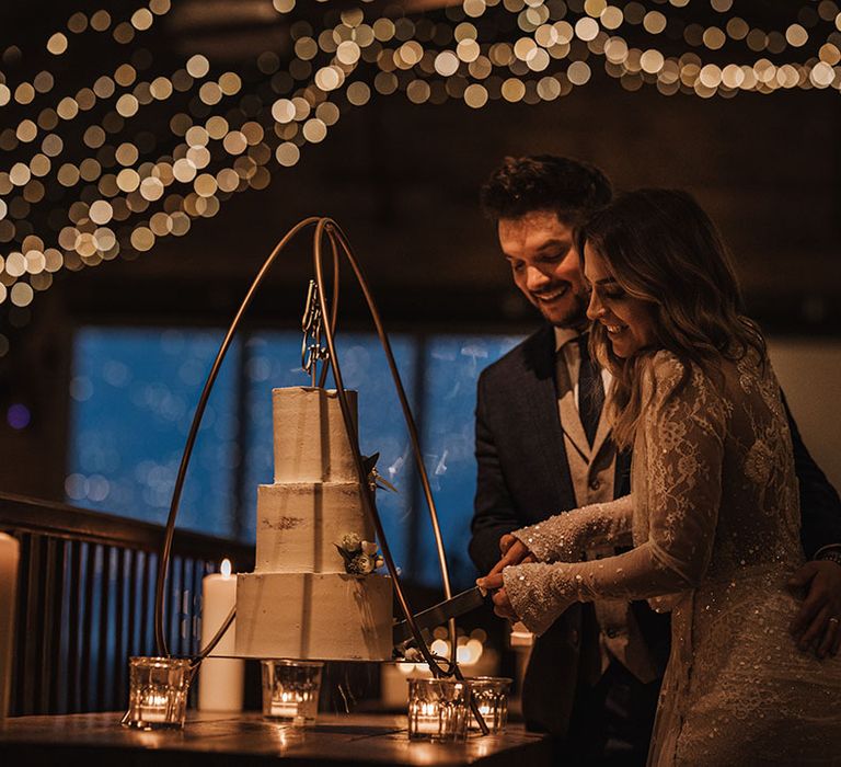 The bride in sequinned long sleeve wedding dress cuts the three tier white iced wedding cake with the groom