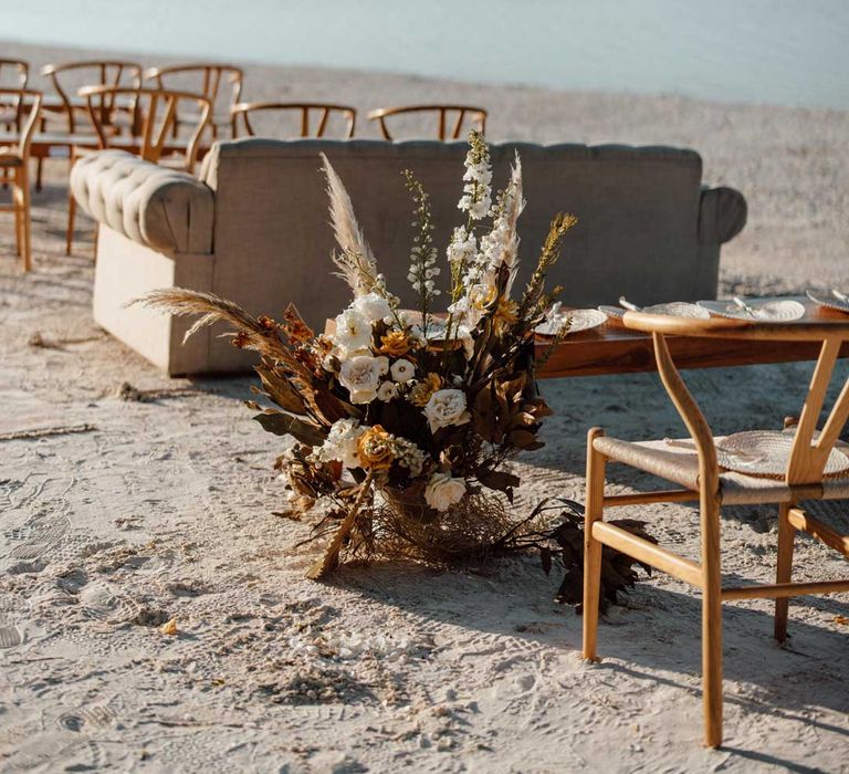 White sofa and wooden chairs on beach wedding with pampas grass, burnt orange dried flower, white garden rose and peony flower arrangements 