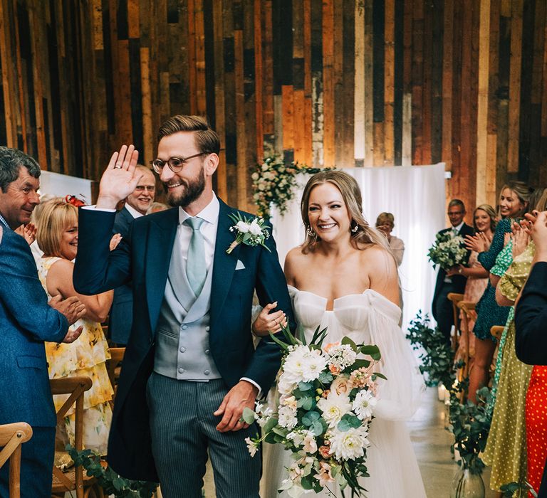 Bride holding a pink and white cascading bouquet in long sleeve wedding dress walking with the groom in a navy suit 