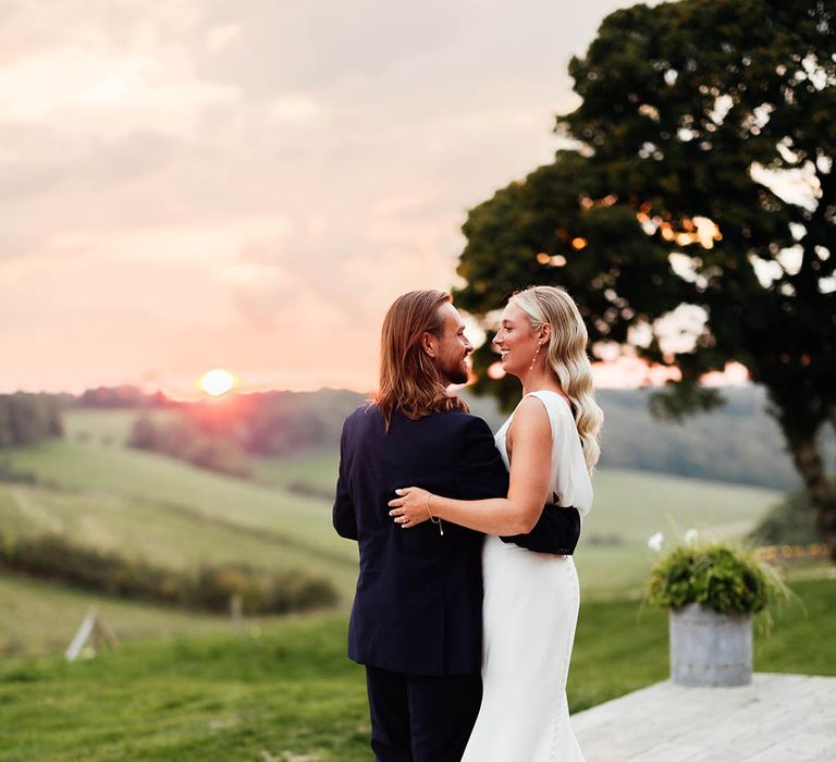 Gorgeous sunset wedding photography with the bride in a draped back wedding dress with a train embracing the groom in a blue suit 