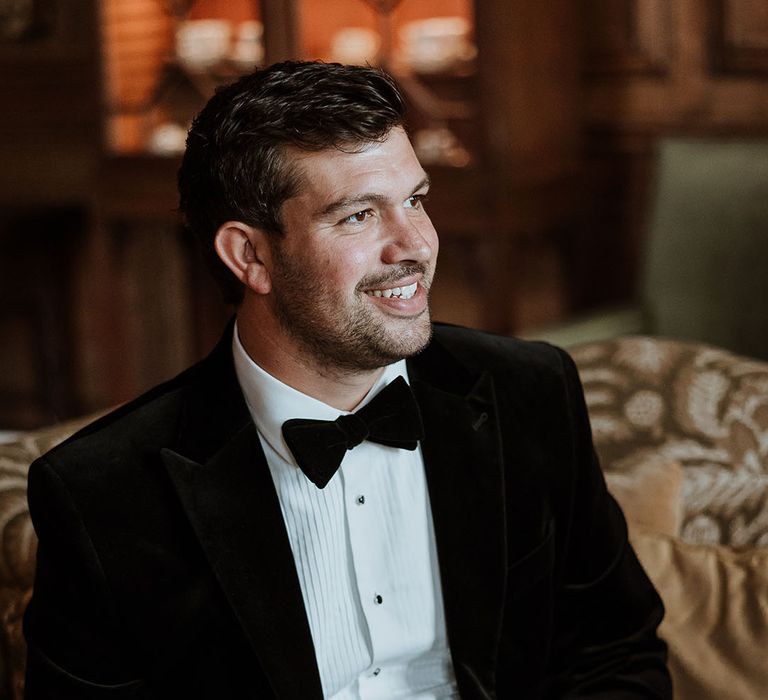 Groom sits wearing a black velvet tuxedo with a velvet bow tie drinking glass of champagne 