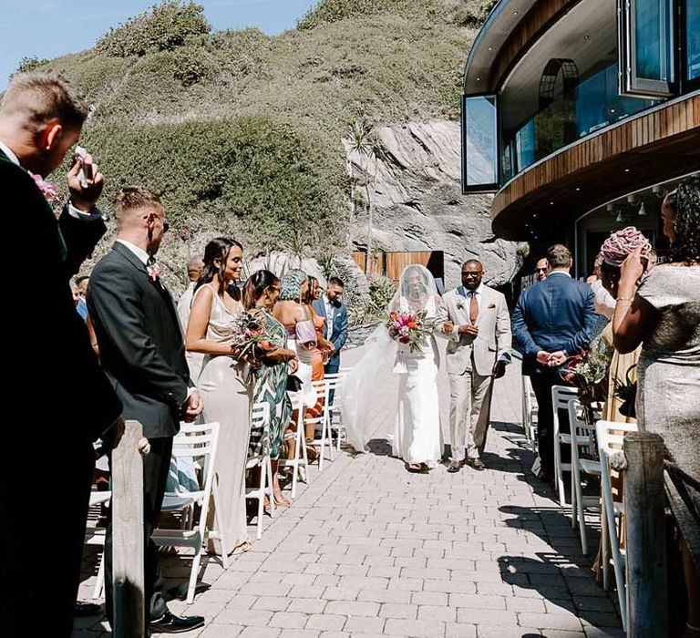 Father of the bride in light grey suit walking down the aisle arm in arm with bride in v neck sleeveless lace Pronovias wedding dress, cathedral length veil and silver sparkly bridal jewellery 