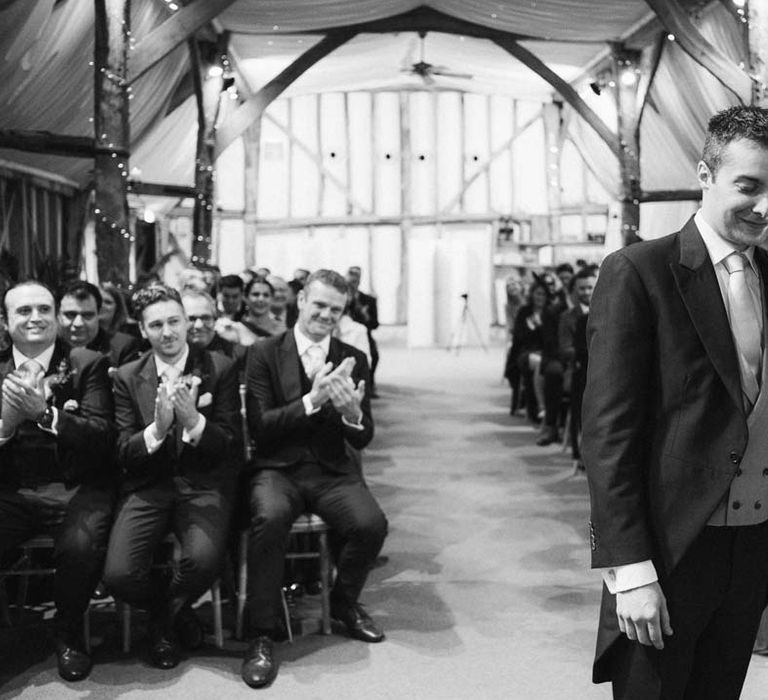 Groom in navy grooms morning suit with grey waistcoat, off-white tie and mixed wildflower boutonniere standing with bride in a-line tulle wedding dress with beaded lace applique with petals and a square neckline with side tulle cutouts at the alter of South Farm wedding venue