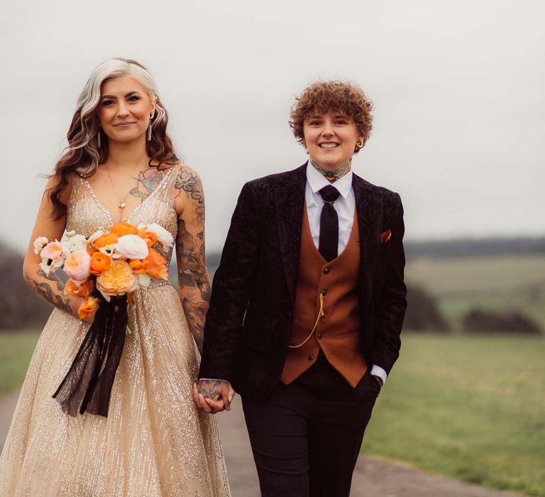 Bride in sleeveless deep v-neck sparkly gold wedding dress holding orange garden rose, white poppy, light pink carnation and foliage bridal bouquet wrapped in sparkly gold ribbon with bride in dark velvet bridal jacket with rust waistcoat and pocket square, black suit trousers and gold pocket watch at Cotswolds wedding venue